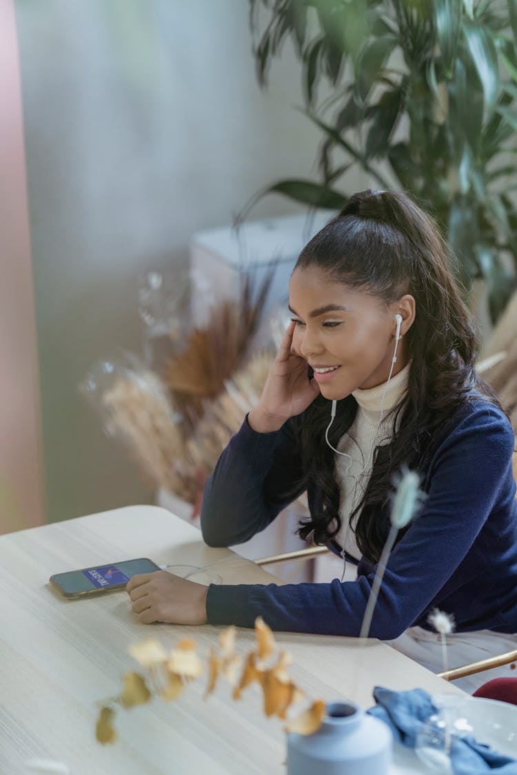 Delighted Young Black Woman In Earphones Sitting At Table And Having Phone Call