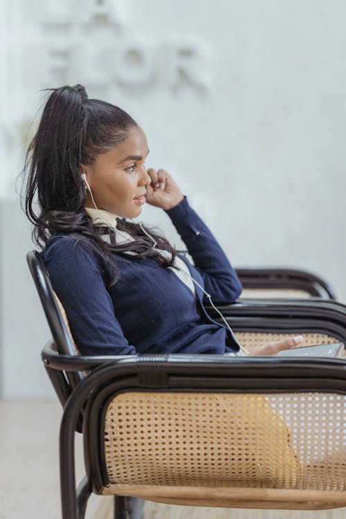 Side view of concentrated young African American businesswoman with long dark hair in elegant clothes using earphone while having phone call sitting in armchair in light office