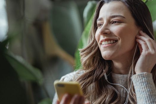 Happy young woman with long wavy hair smiling and adjusting earphones while listening to music via mobile phone