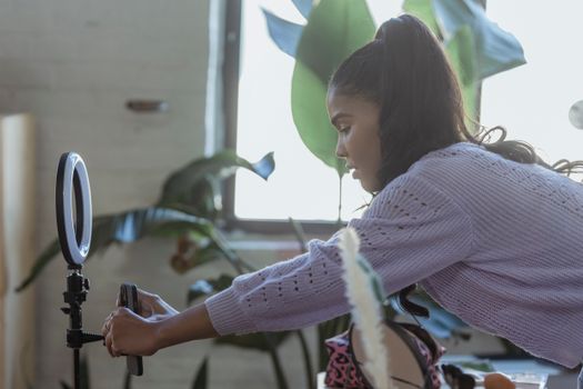 Side view of concentrated young African American female blogger with long dark hair in stylish sweater attaching smartphone to tripod with ring light before recording vlog
