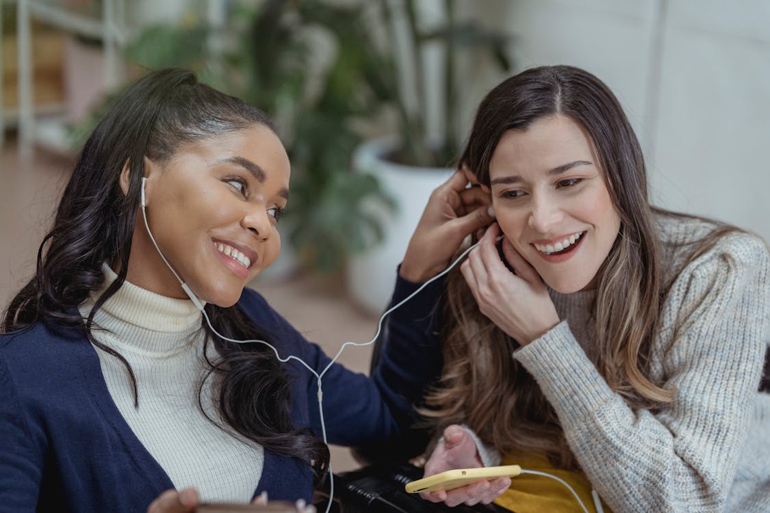 digital . Free Happy young African American lady with smartphone and earphones smiling and sharing music with female best friend while chilling together in cozy cafe Stock Photo