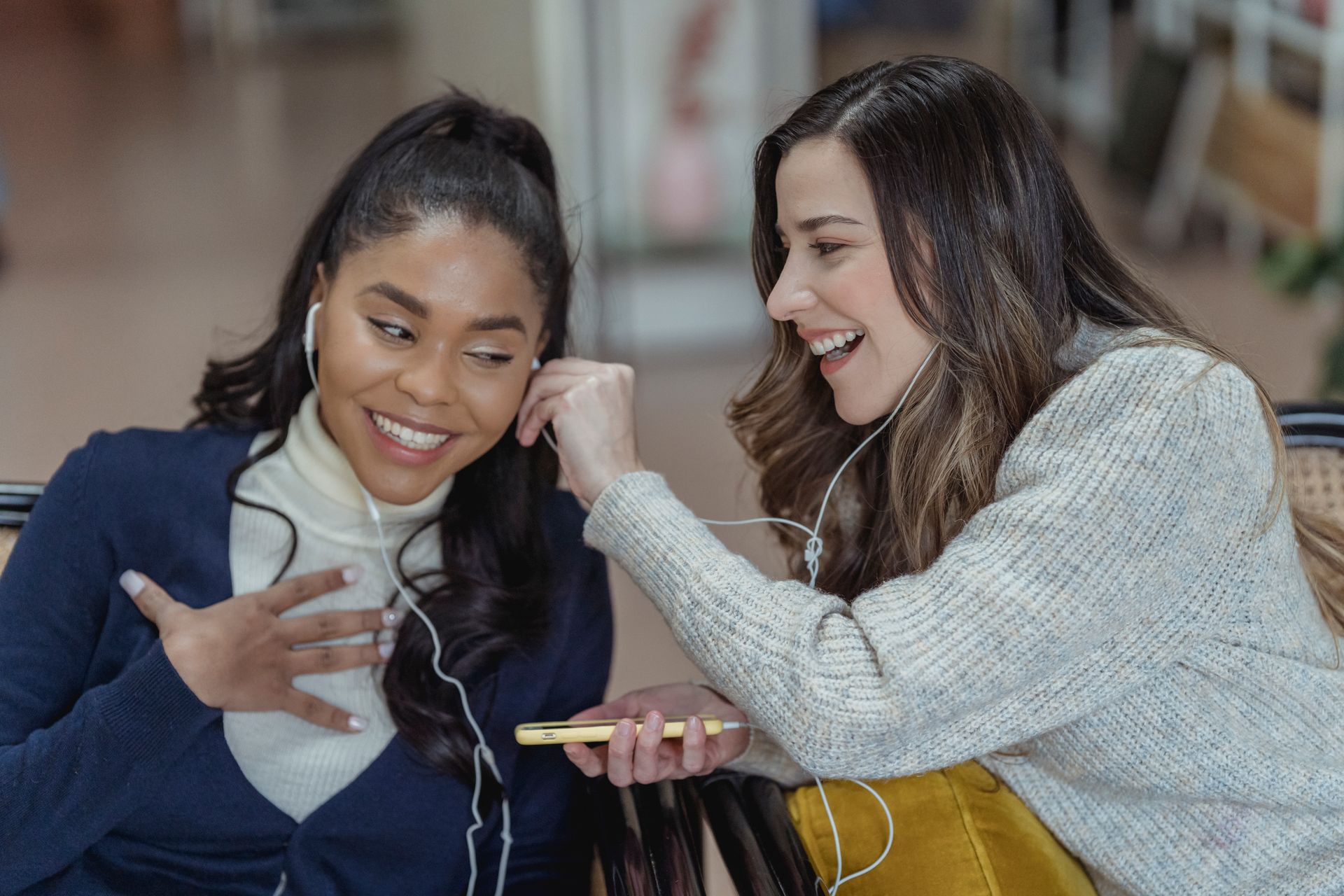 Multiracial cheerful women smiling and enjoying music in earphones while using mobile phone on blurred background