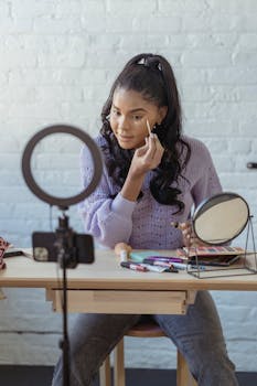 Young African American female blogger spreading foundation cream on face at table with cosmetics near smartphone recording video