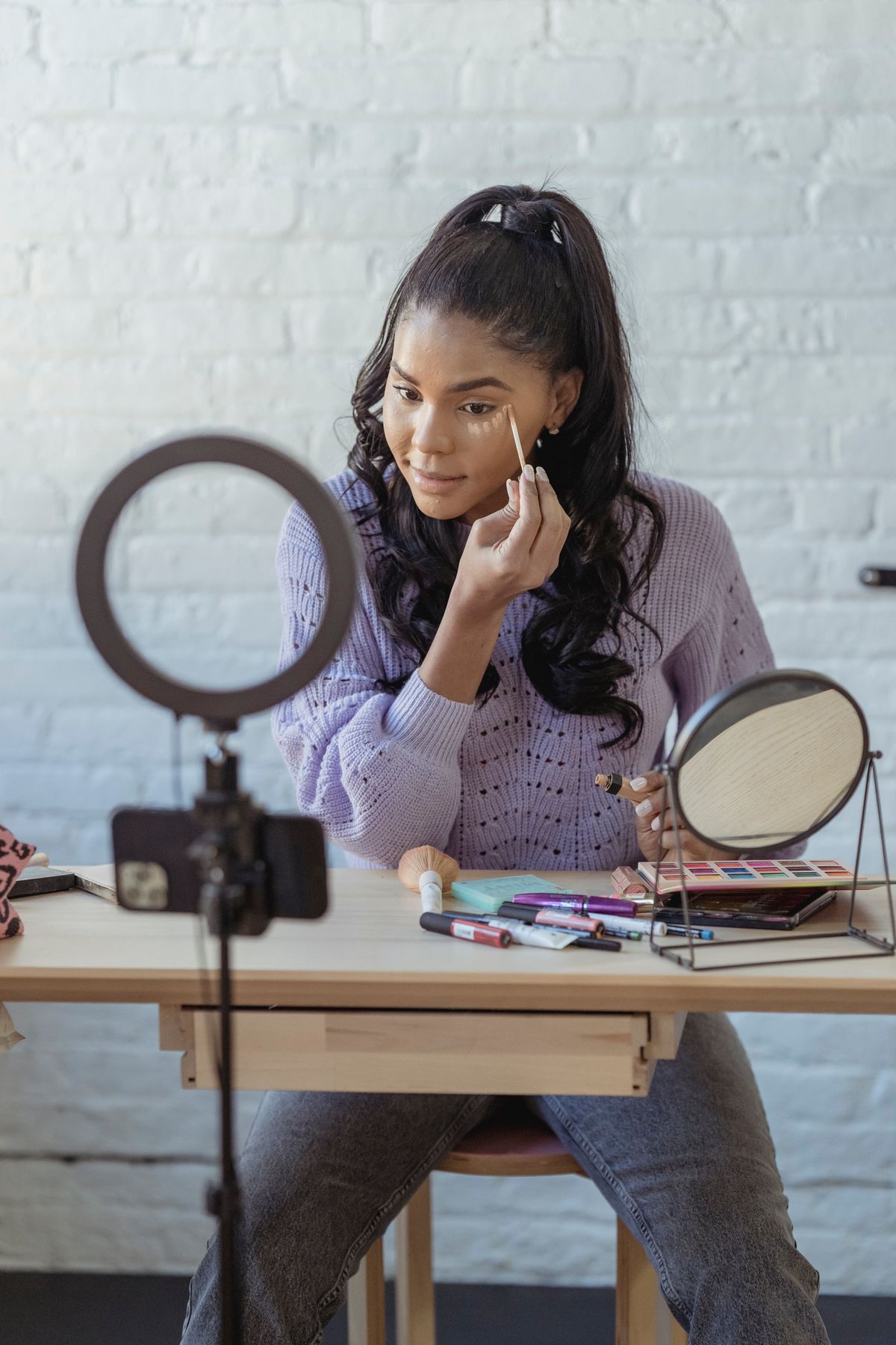 A Black woman recording a beauty tutorial with a smartphone beside a display of makeup products.
