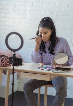 Serious stylish African American female beautician spreading foundation on face with soft professional cosmetic brush during online session