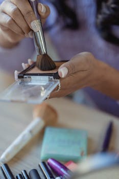 Crop unrecognizable ethnic woman sitting at table and using brush to take powder while applying makeup in light room