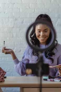 Smiling African American female applying makeup and filming vlog on smartphone in light room in daytime
