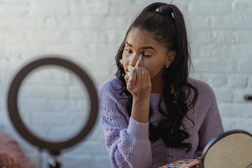 Concentrate African American female with hairstyle applying eyeliner while making makeup in light room against brick wall