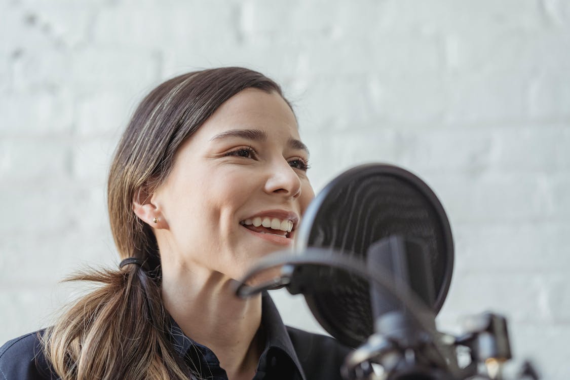 Positive female recording podcast on mic while working in light studio against white brick wall