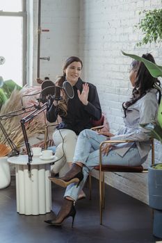 Full body of female journalist asking question to African American woman sitting in armchair with crossed legs