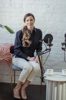 Full body of positive female host wearing formal clothes sitting in armchair near table with cups of coffee and microphone for interview