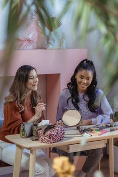 Positive multiethnic female bloggers sitting at table with cosmetic products and mirror and recording vlog on cellphone on tripod