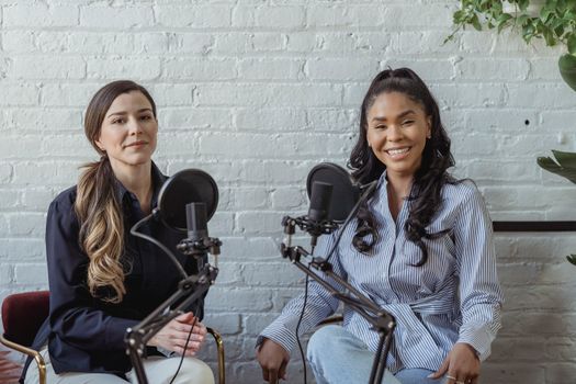 Smiling African American female guest sitting near radio host at interview against white brick wall and looking at camera