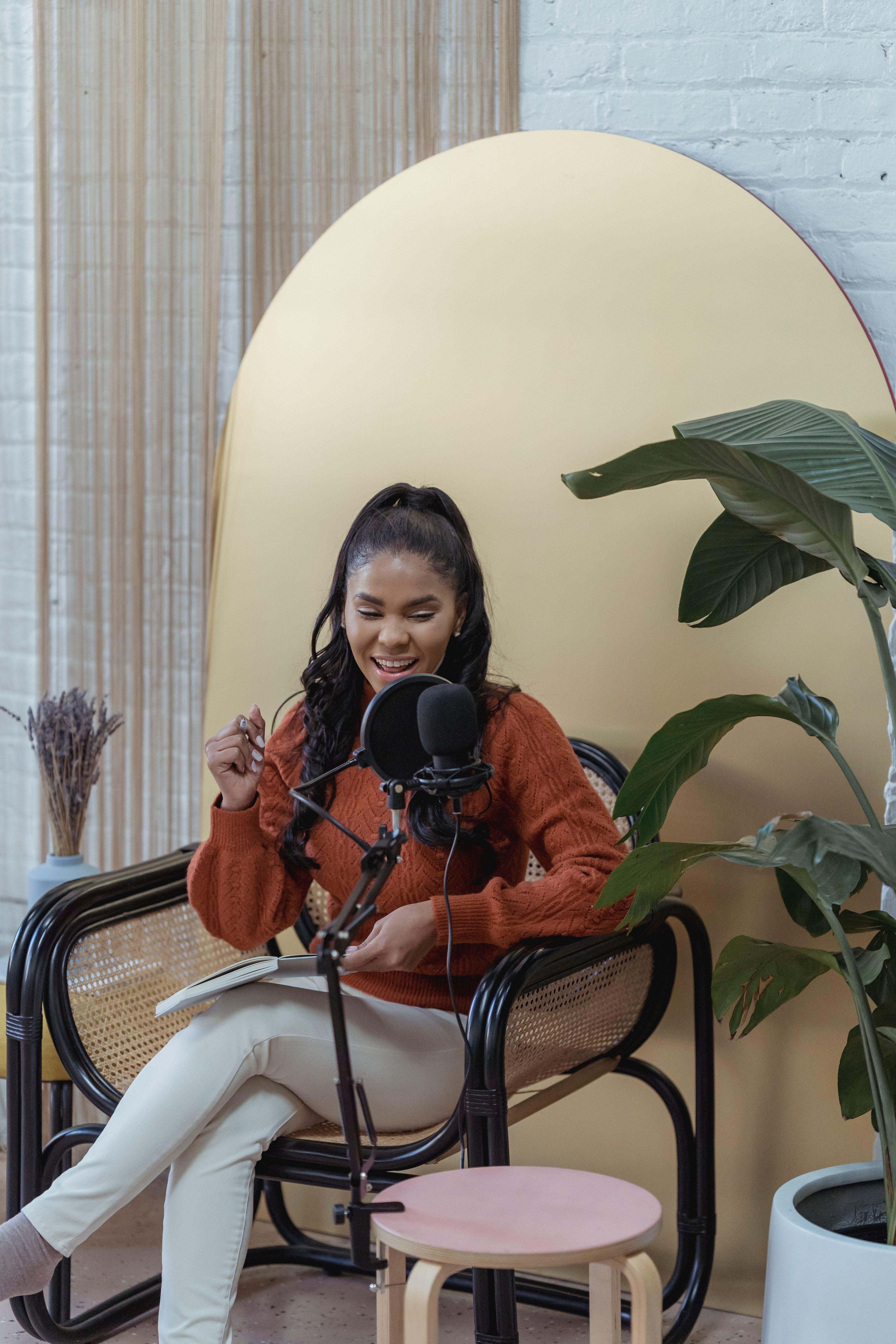 African American woman joyfully recording a podcast in a cozy, stylish indoor setting.