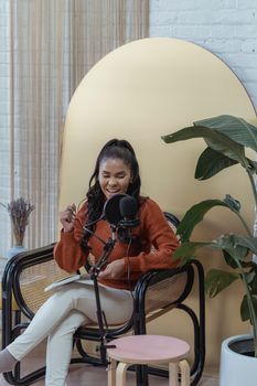 Delighted African American female sitting in armchair and talking to microphone while sitting with crossed legs with notebook