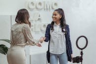 Positive African American female shaking hand of partner standing in light office near smartphone with ring lamp on tripod