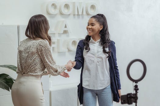 Positive African American female shaking hand of partner standing in light office near smartphone with ring lamp on tripod