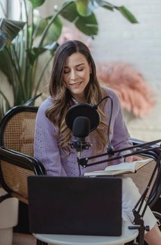 High angle of positive female radio host sitting in armchair and using netbook and mic for remote work