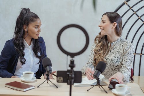 Free Positive blogger asking questions to African American speaker and recording interview on cellphone at table with microphones Stock Photo