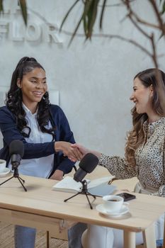 Smiling multiethnic women sitting at table with microphones and cups of coffee near smartphone and greeting each other with handshake during interview