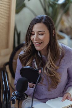 Positive female radio host with professional microphone sitting on chair while recording audio message in light room with green plants