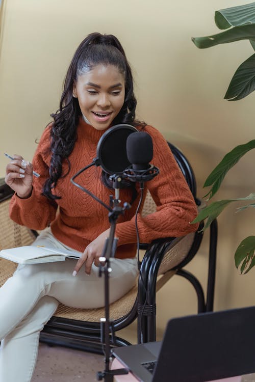 Positive African American female recording podcast with microphone while sitting in chair with notebook in light room with modern laptop