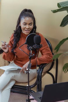 Positive African American female recording podcast with microphone while sitting in chair with notebook in light room with modern laptop