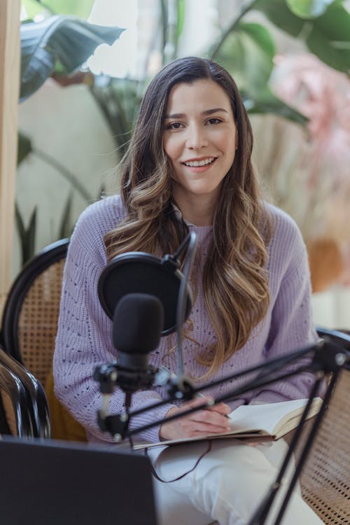 Free Cheerful female radio host looking at camera while sitting in chair with opened notebook near professional microphone in light room Stock Photo