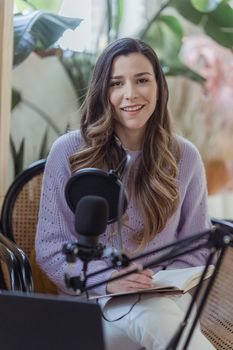 Cheerful female radio host looking at camera while sitting in chair with opened notebook near professional microphone in light room
