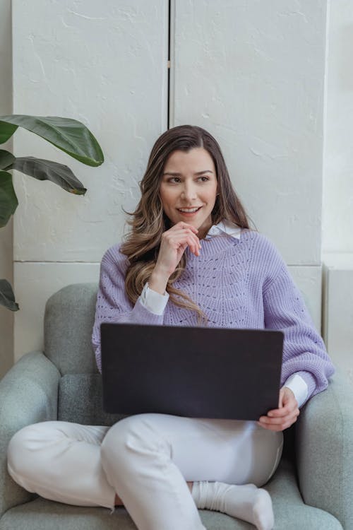 Cheerful female worker surfing internet on modern netbook and looking away while sitting in comfortable armchair in light room during remote work