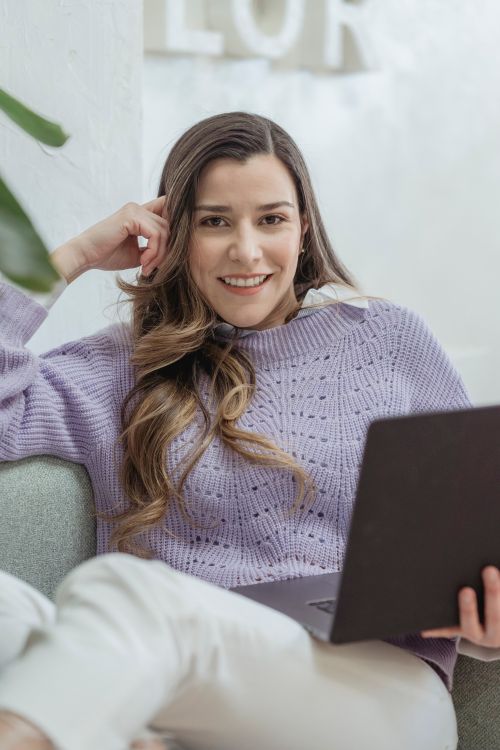 Free Happy young female in violet sweater holding netbook and sitting on cozy couch while looking at camera with smile Stock Photo