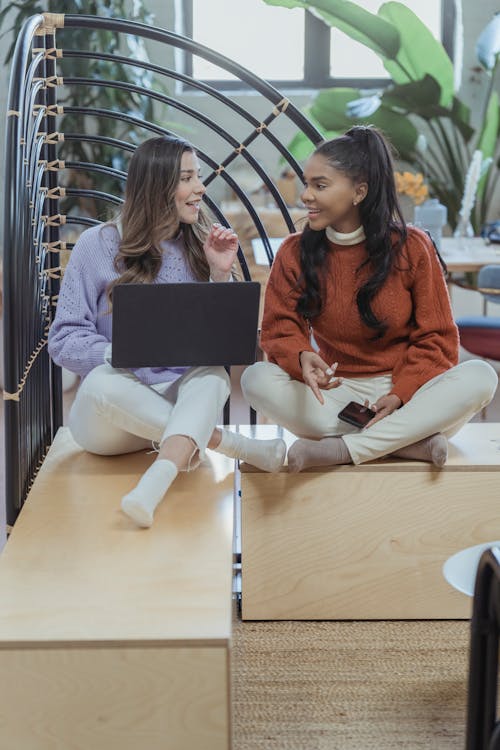 Full body content multiethnic female friends in casual outfits browsing netbook and sharing ideas while sitting together with legs crossed