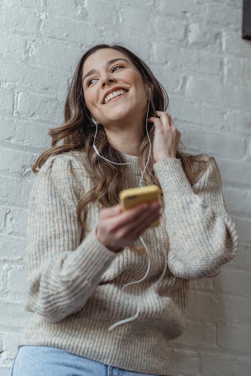 Free Cheerful young female in sweater browsing mobile phone and listening to favorite music in earphones against brick wall Stock Photo