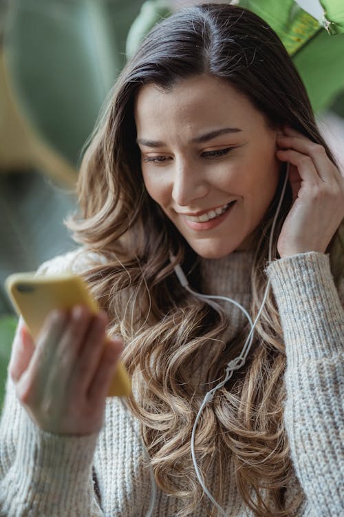 Free Young content female in casual sweater browsing modern mobile phone and listening to good music via earphones in living room Stock Photo