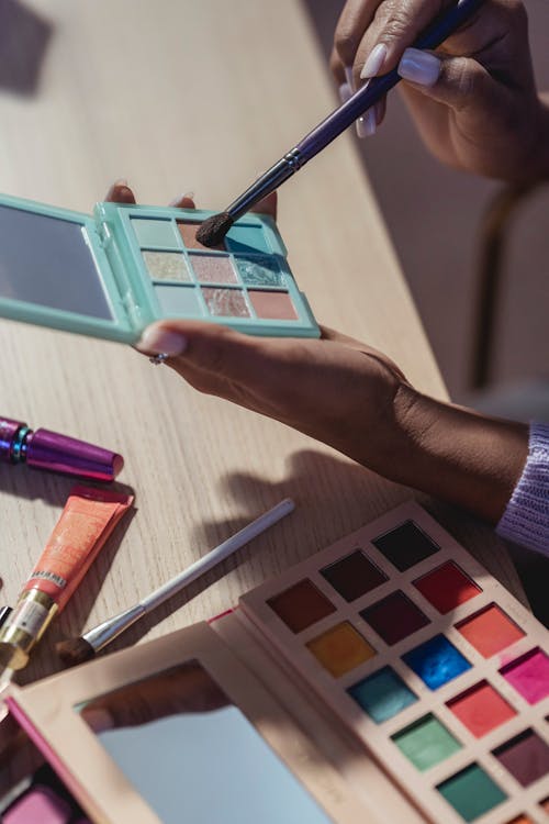 Crop anonymous African American female visagiste applying shiny eyeshadow on brush while sitting at table with makeup products