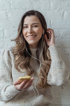 Young content female in sweater and earphones browsing mobile phone and looking at camera while standing against brick wall and touching hair