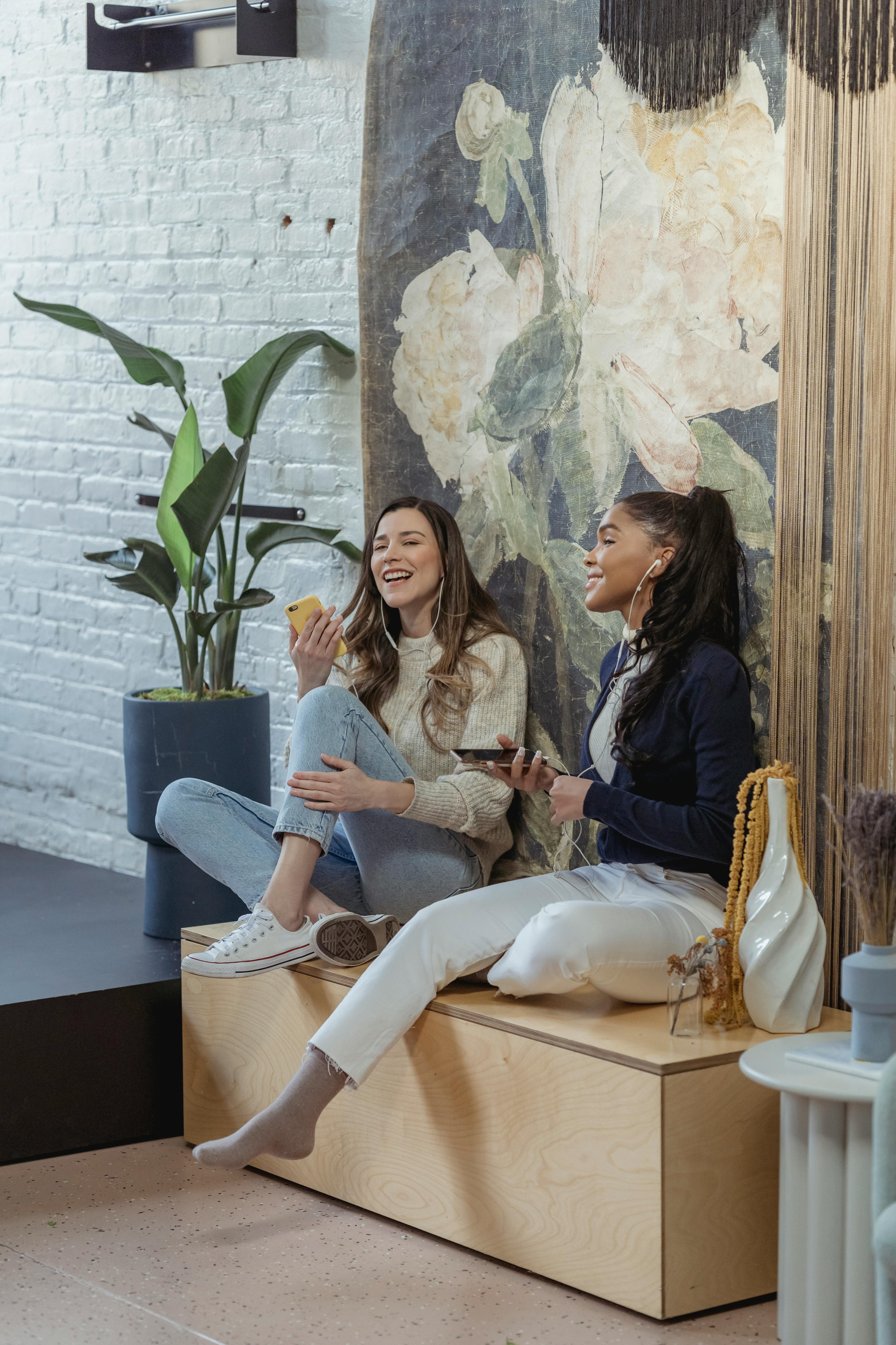 Full body joyful multiracial female friends in casual clothes using smartphones and listening to good music via earphones in modern living room