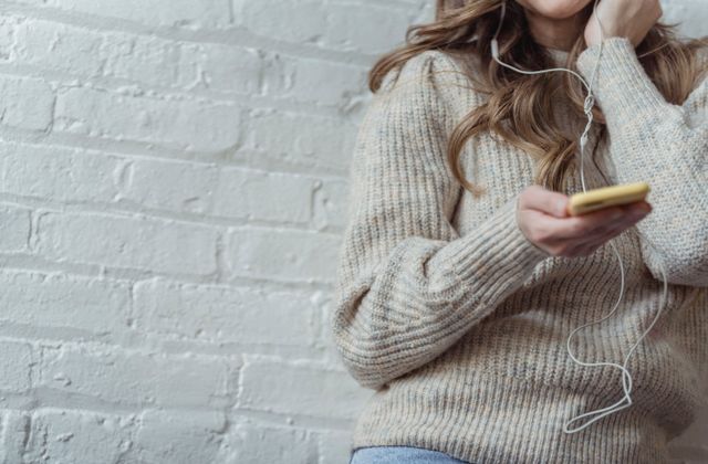 A person in a warm sweater listening to music through earphones on their phone