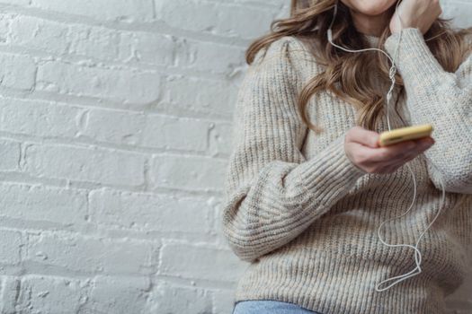 Crop unrecognizable female with long wavy hair in warm sweater listening to music in earphones via mobile phone while sitting near white brick wall
