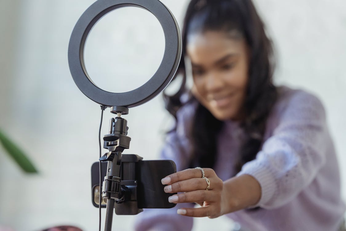 Cheerful young African American female blogger in stylish sweater smiling while setting up camera of smartphone attached to tripod with ring light before recording vlog
