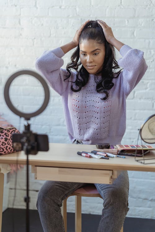 Free Stylish young African American female influencer in sweater adjusting long dark hair while sitting at wooden table before recording makeup tutorial video on smartphone Stock Photo