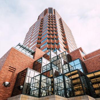 From below of multistory contemporary multistage building with geometric facade made of stone bricks and metal panels with trees reflecting in glass windows under cloudy sky on sunny day