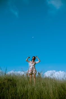 Distant female in dress with raised arms standing on grassy field while admiring nature on sunny summer day in countryside