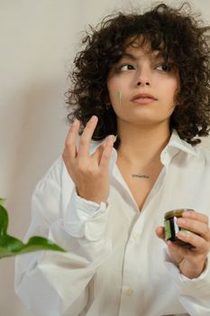 A woman with curly hair applies face cream, emphasizing skincare and self-care in a cozy indoor setting.