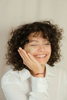Young woman with curly hair, eyes closed, enjoying skincare routine indoors.