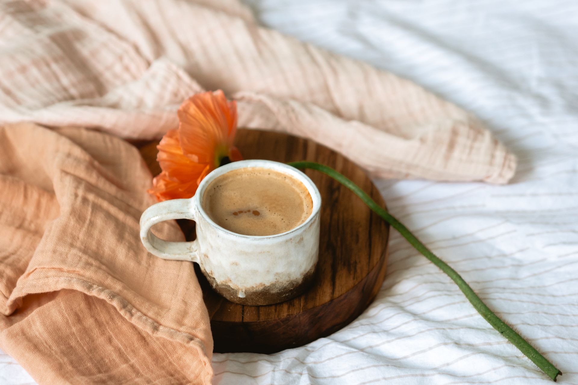 A warm coffee cup placed on a wooden tray adorned with an orange flower, wrapped in soft fabric.