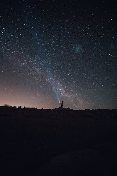 Breathtaking view silhouette of anonymous person standing on ground and enjoying view of starry sky while lighting blue beam to dark night at sunset
