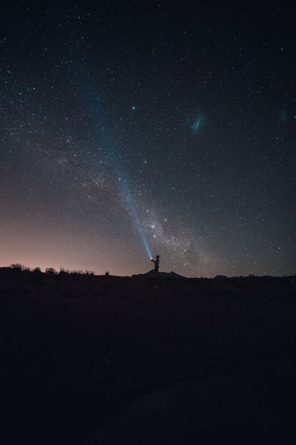 Breathtaking view silhouette of anonymous person standing on ground and enjoying view of starry sky while lighting blue beam to dark night at sunset