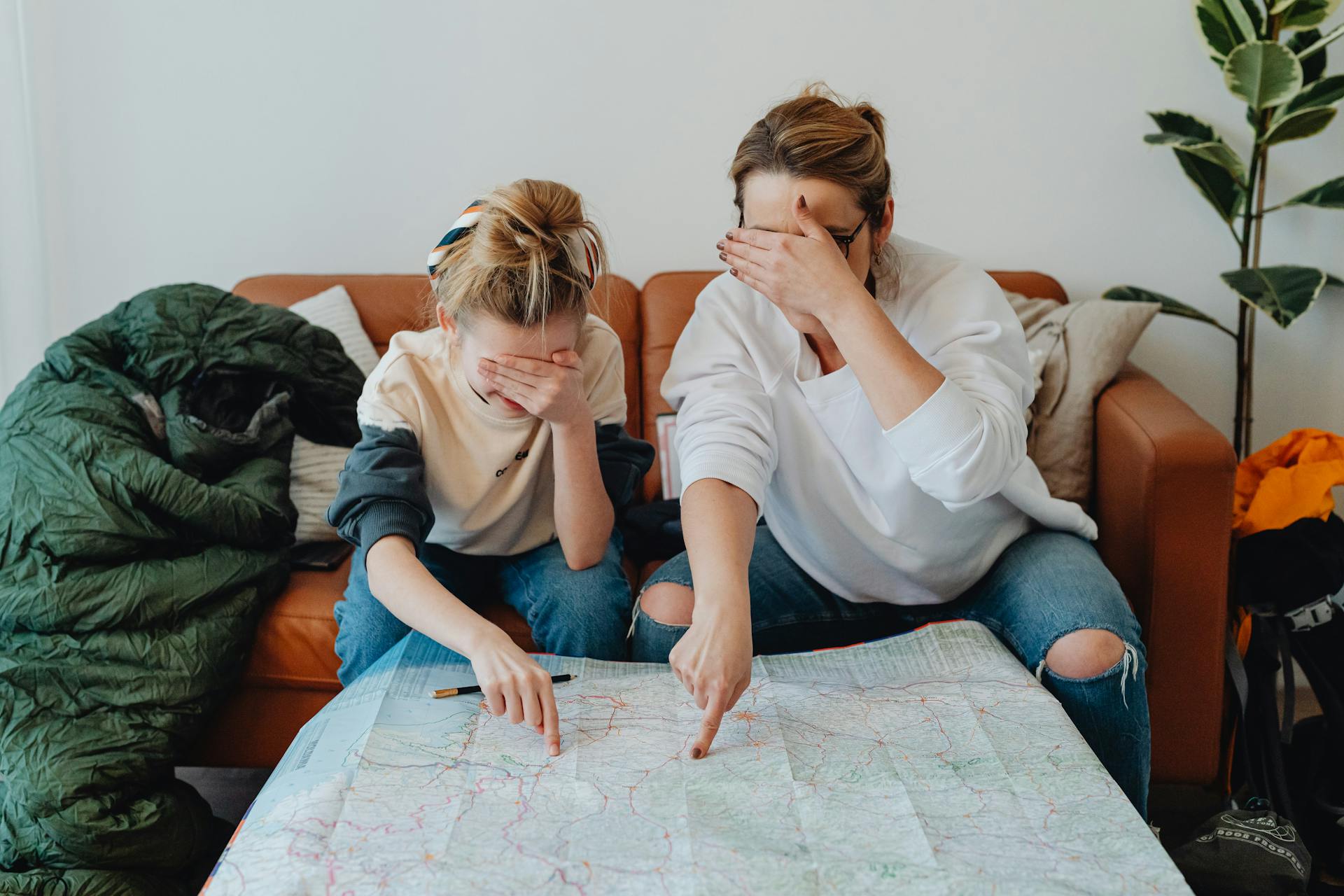 Mother and daughter covering eyes while pointing at map on couch, planning a trip.