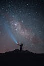 在无云的星空下，一名匿名游客的剪影站在悬崖上，用手电筒发出信号